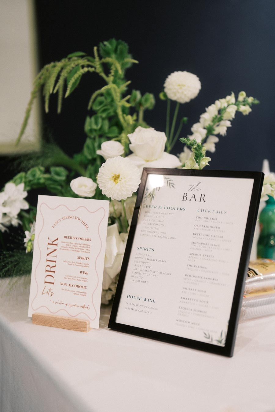 Elegant bar setup featuring a floral arrangement and two printed drink menus on a white tablecloth background; perfect for a wedding or formal event.