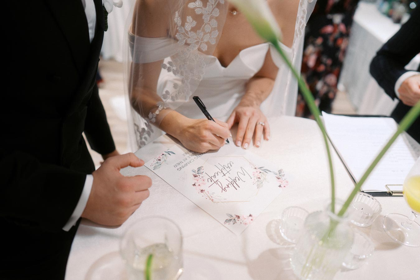 Bride signing a wedding certificate at a decorated table, accompanied by the groom in a formal suit.