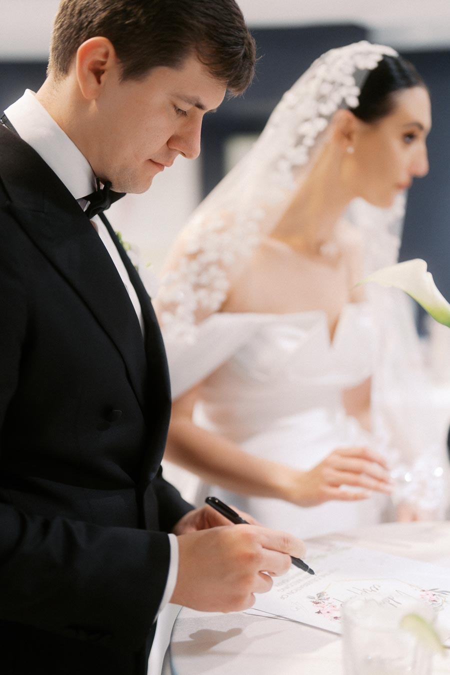A groom in a black tuxedo and a bride in an elegant white dress and veil, signing their wedding certificate at the ceremony.
