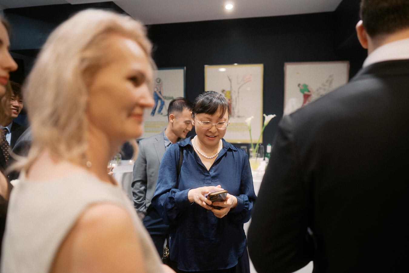 A group of people socializing at an indoor event, with a woman in glasses and a pearl necklace looking at her phone in the center.