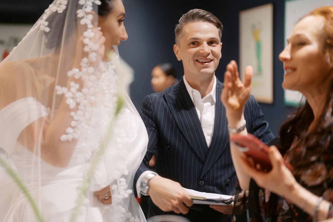 Wedding scene with a bride in a floral veil talking to a smiling man in a pinstripe suit, with a woman gesturing nearby.