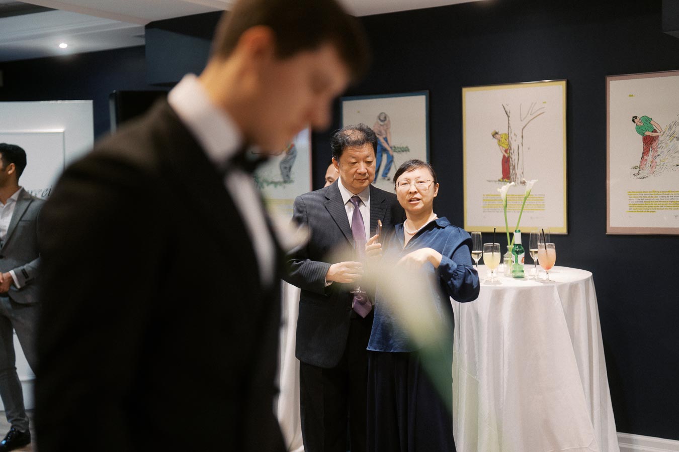 A couple engaging in conversation at an art gallery event, with framed artwork on a dark wall in the background and a table adorned with drinks and flowers in the foreground.