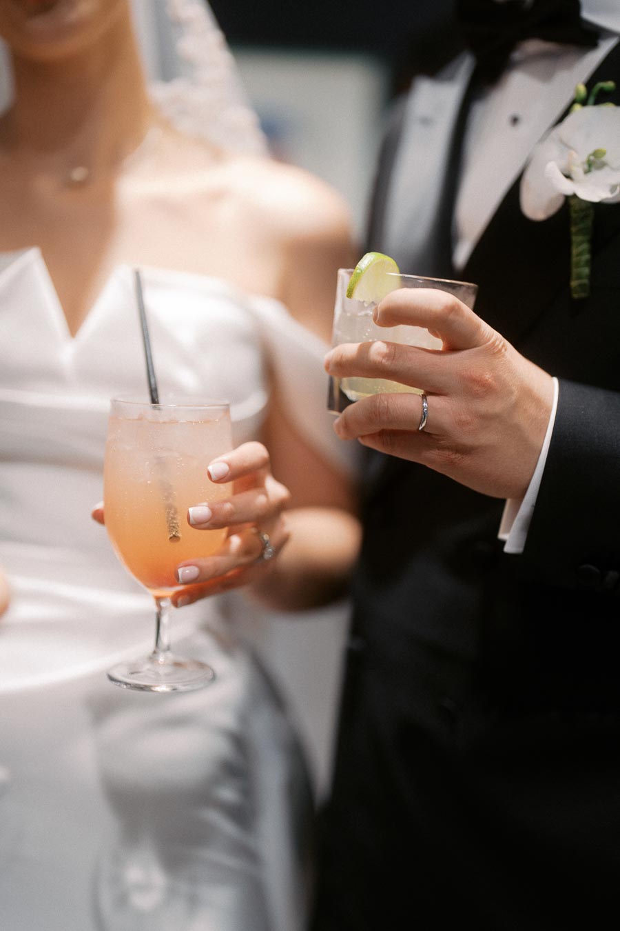 Bride and groom toasting with cocktails at a wedding reception, featuring elegant attire and close-up view of rings and drinks.