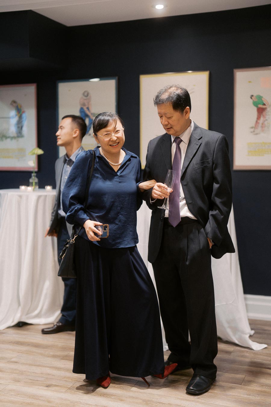 Two people dressed in formal attire enjoying a conversation at a social event, with framed artwork on the wall and a table with white cloth in the background.