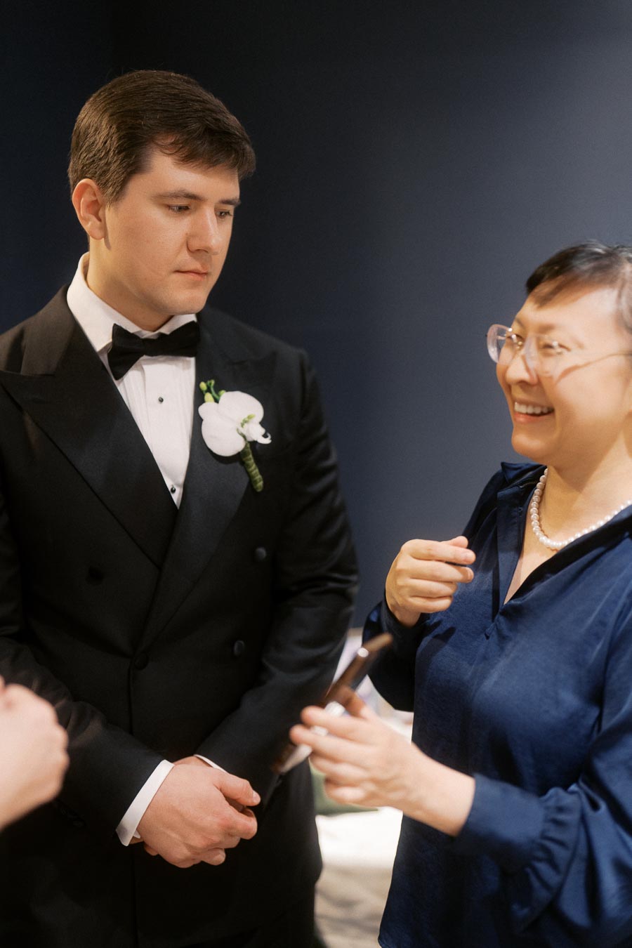 A well-dressed man in a black tuxedo with a white boutonniere is engaged in conversation with a smiling person wearing glasses and a blue blouse, set against a dark background.