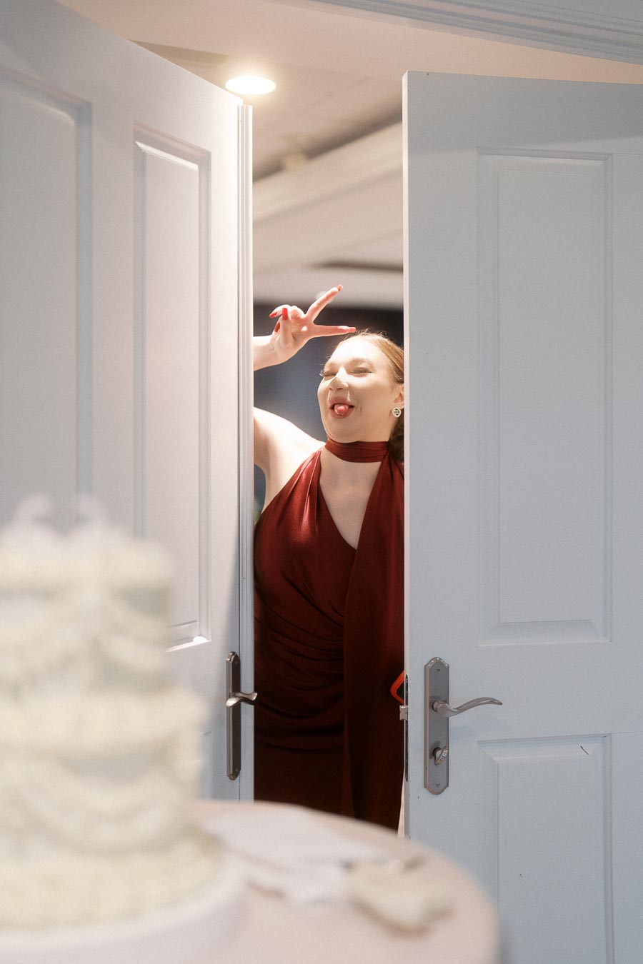 Person playfully posing with a peace sign and tongue out in a doorway, wearing a red dress, with a blurred view of a white cake in the foreground.