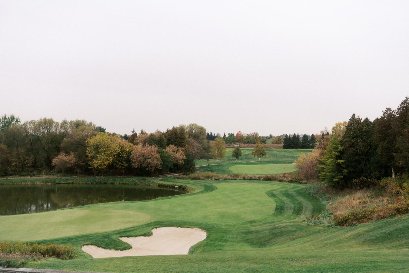 Scenic view of a well-maintained golf course featuring lush green fairways, a sand bunker, and a pond, surrounded by autumn trees under a clear sky. Ideal for outdoor enthusiasts and golf lovers.