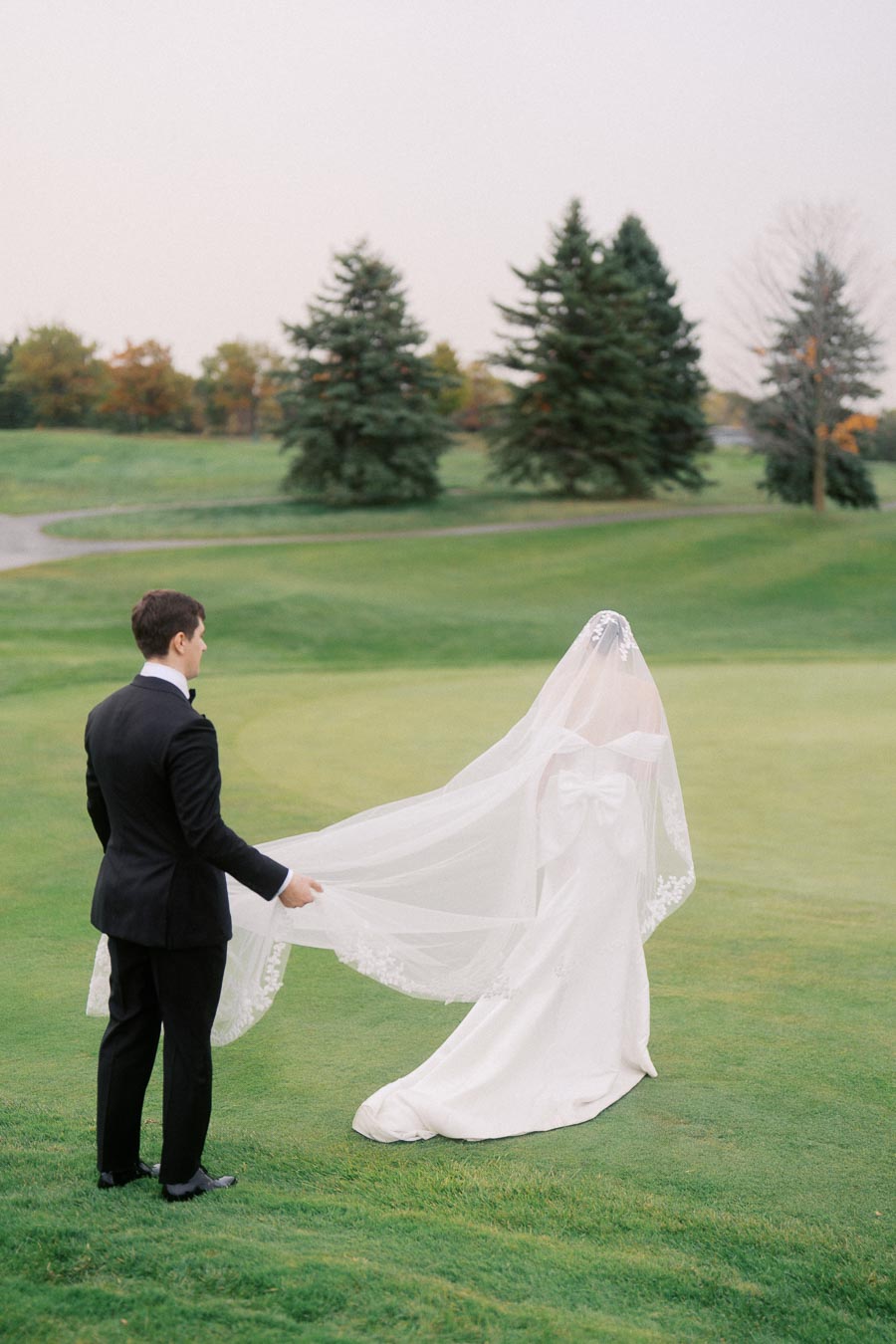 A bride in an elegant white wedding dress with a long veil stands on a lush green lawn, as the groom gently holds the veil. The romantic outdoor setting features a backdrop of evergreen trees under a clear sky.