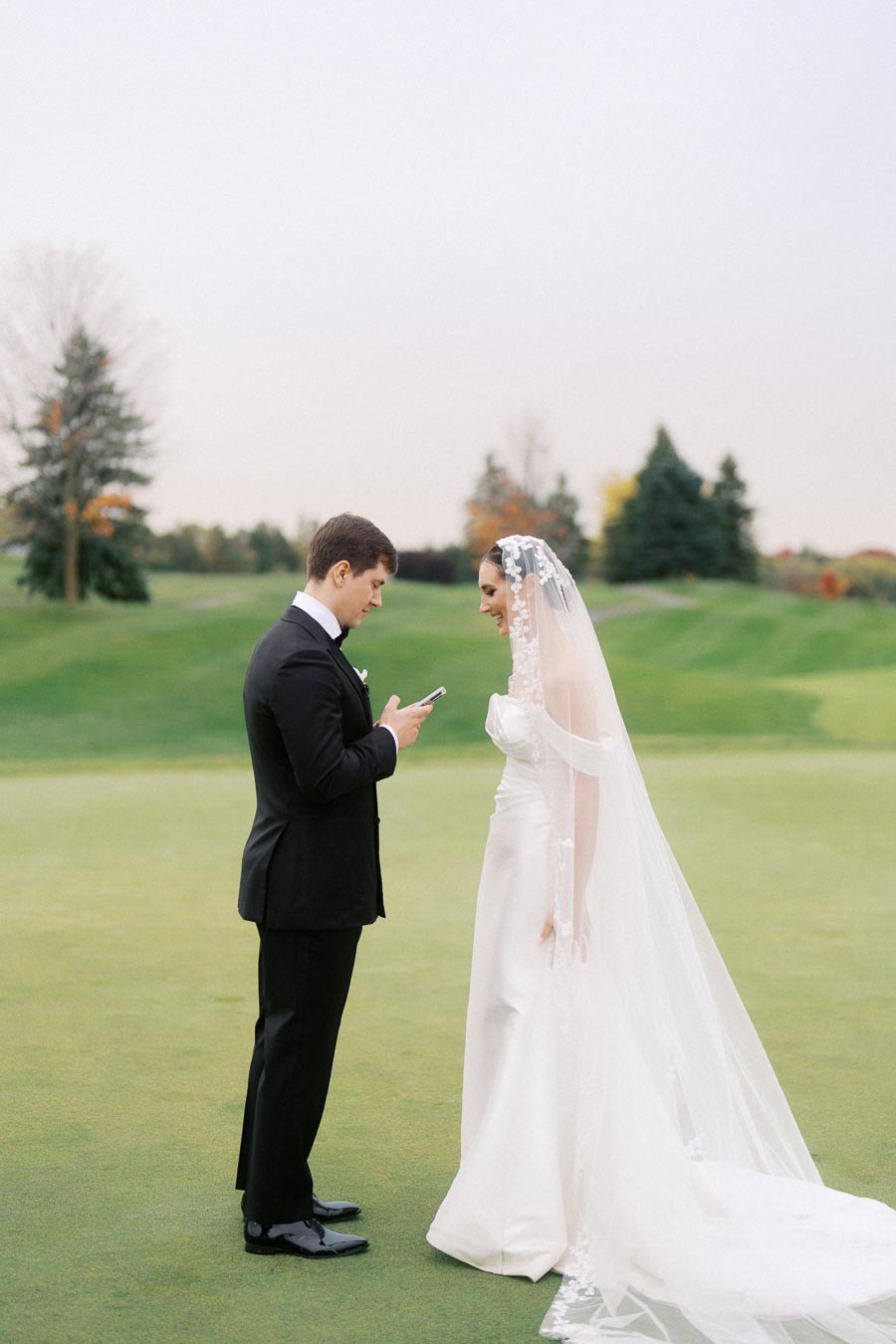 Bride and groom exchanging vows in an outdoor wedding ceremony, with groom reading from phone and bride smiling in elegant white dress and veil, set against a green landscape with trees.