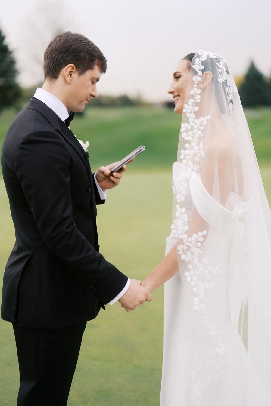 A bride and groom standing outdoors, holding hands while the groom reads vows from a phone. The bride is wearing a white dress with a floral veil, and the groom is in a black tuxedo.