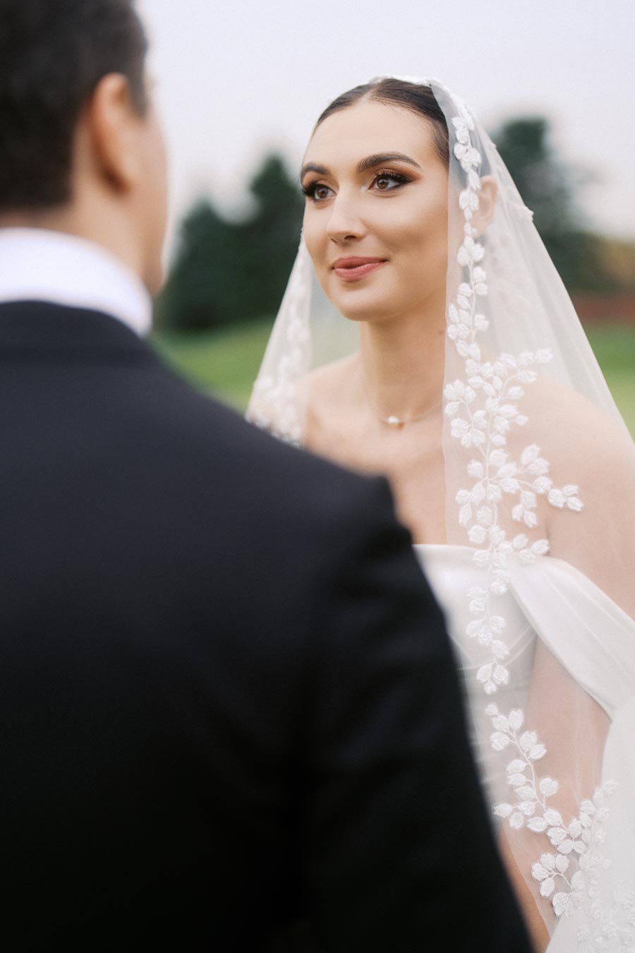 Bride in elegant white gown and floral veil gazes lovingly at groom during outdoor wedding ceremony.