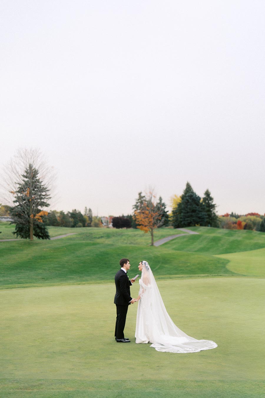 Bride and groom exchanging vows on a golf course, surrounded by lush greenery and autumn trees. The bride is wearing a long white gown with a veil, and the groom is in a black suit.