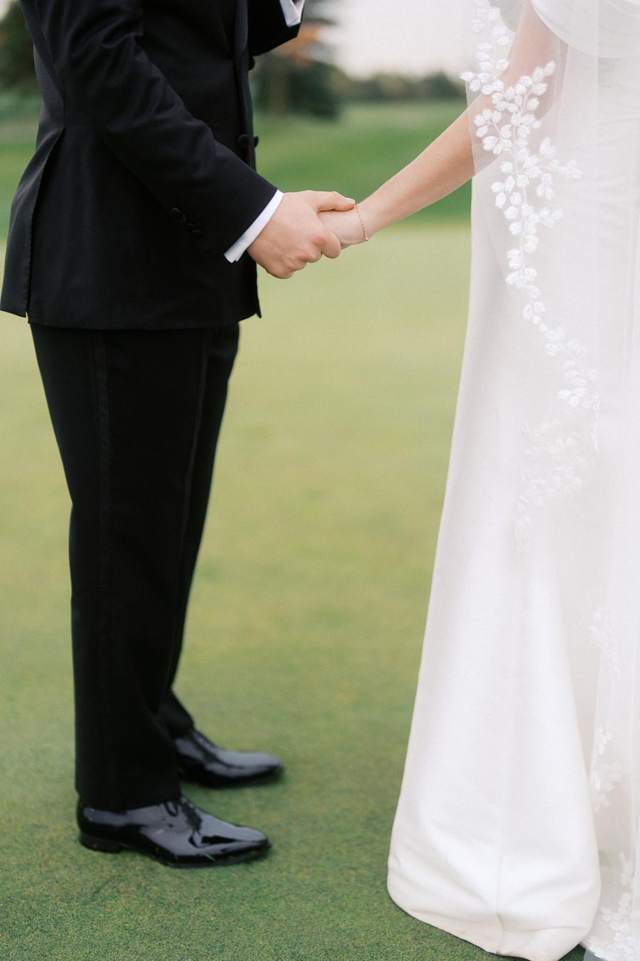 Elegant bride and groom holding hands on a lush green lawn, showcasing wedding attire details.