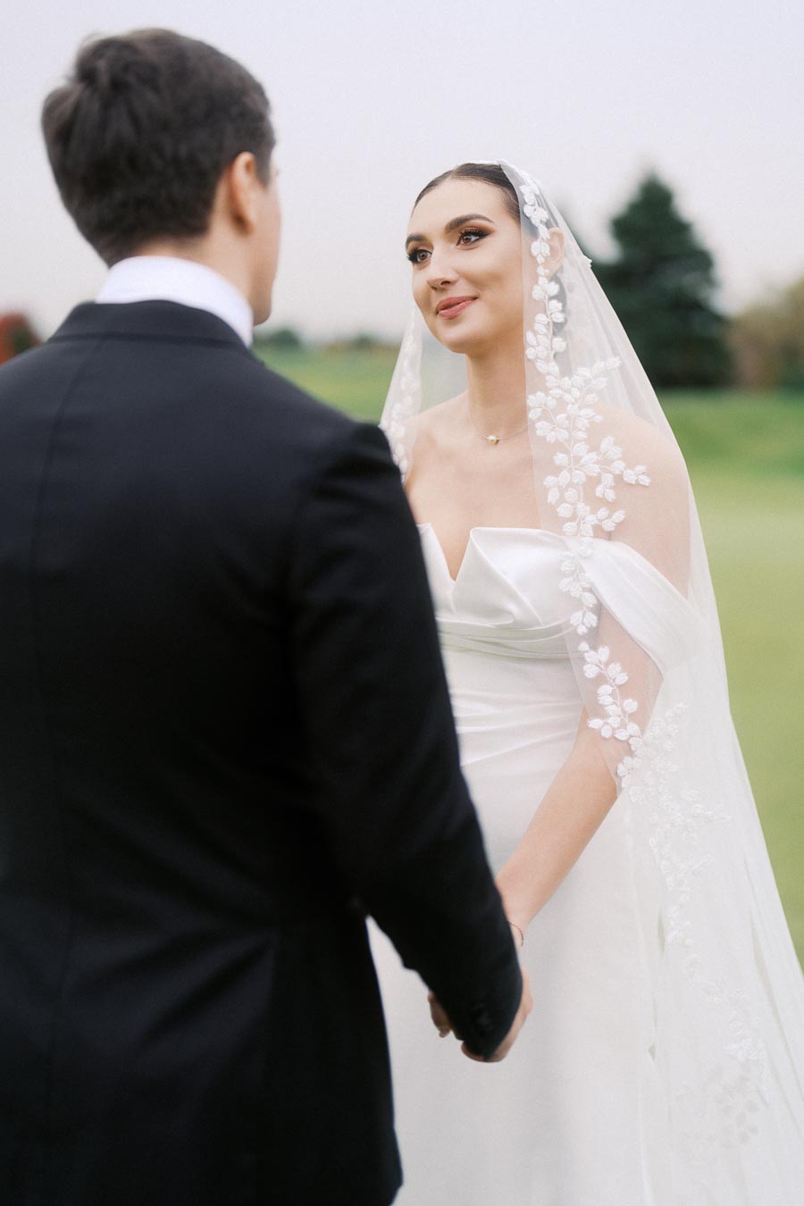 Bride in elegant white gown and lace veil holding hands with groom in black suit during outdoor wedding ceremony.