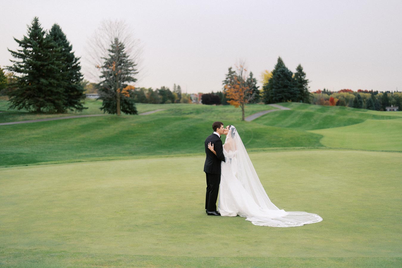 A bride and groom share a kiss on a lush green golf course, surrounded by trees in autumn colors, with the bride wearing a long flowing white veil and gown.