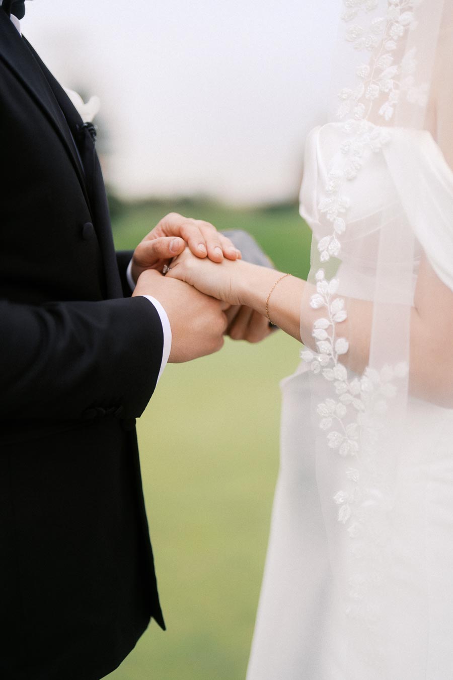 A bride and groom holding hands in an intimate wedding ceremony, with the bride wearing an elegant lace veil and the groom in a classic black suit, set against a serene outdoor backdrop.