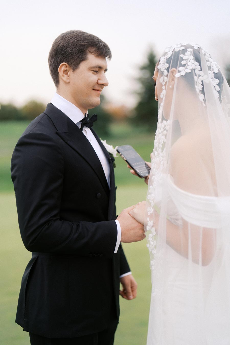 A bride and groom hold hands while exchanging vows during an outdoor wedding ceremony. The groom wears a classic black tuxedo, and the bride wears a veil adorned with floral patterns.