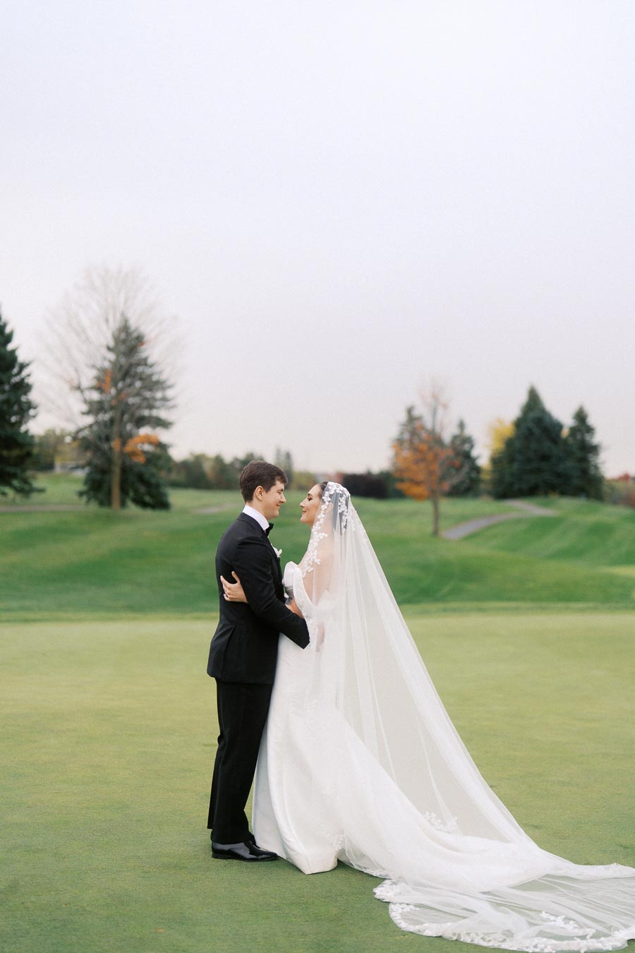 Bride and groom embracing on a golf course, with the bride in a long white gown and veil, and the groom in a black tuxedo, set against a backdrop of autumn trees and a clear sky.