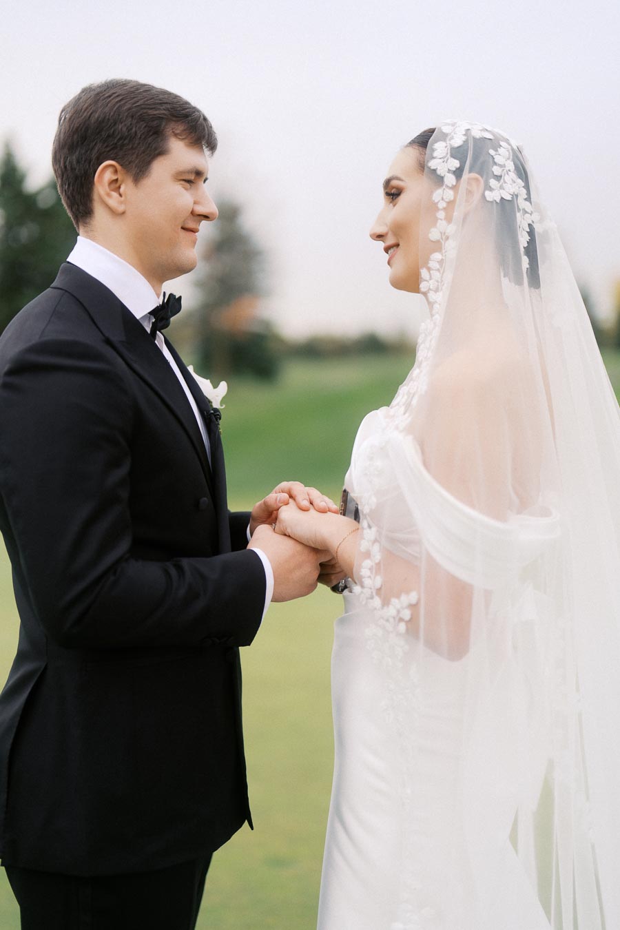 A bride and groom holding hands during an outdoor wedding ceremony, with the bride wearing a lace veil and the groom in a black tuxedo.
