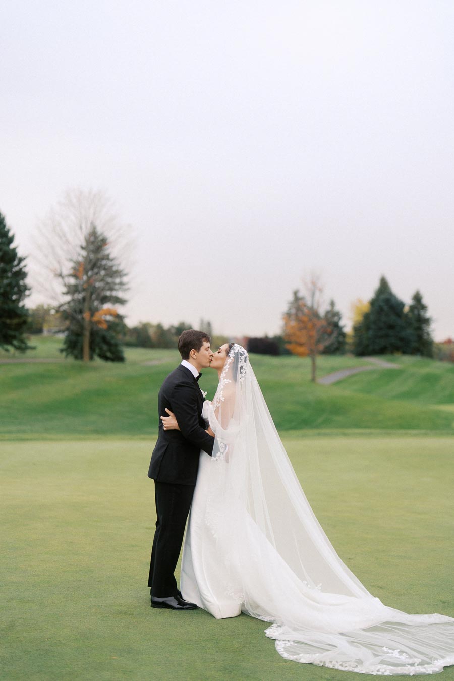 Bride and groom sharing a romantic kiss on a golf course with lush greenery; bride wearing a long, elegant veil and wedding dress, groom in a classic black suit and tie; overcast sky enhancing the serene setting; autumn trees in the background.