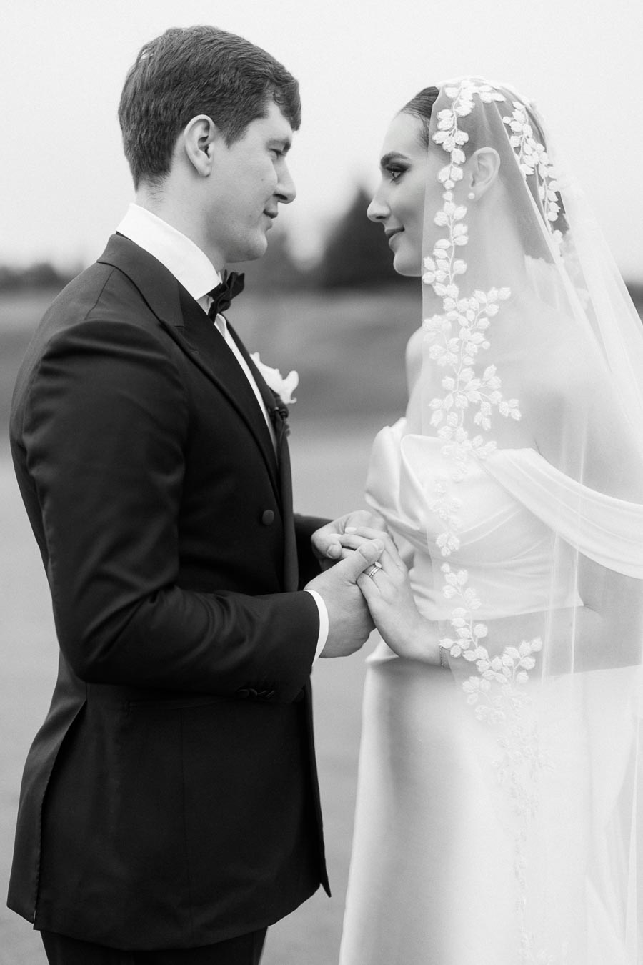 Black and white photo of a bride and groom holding hands, gazing into each other's eyes. The bride wears an embroidered veil over a strapless dress, while the groom is dressed in a formal suit with a bow tie. Romantic wedding moment captured outdoors.