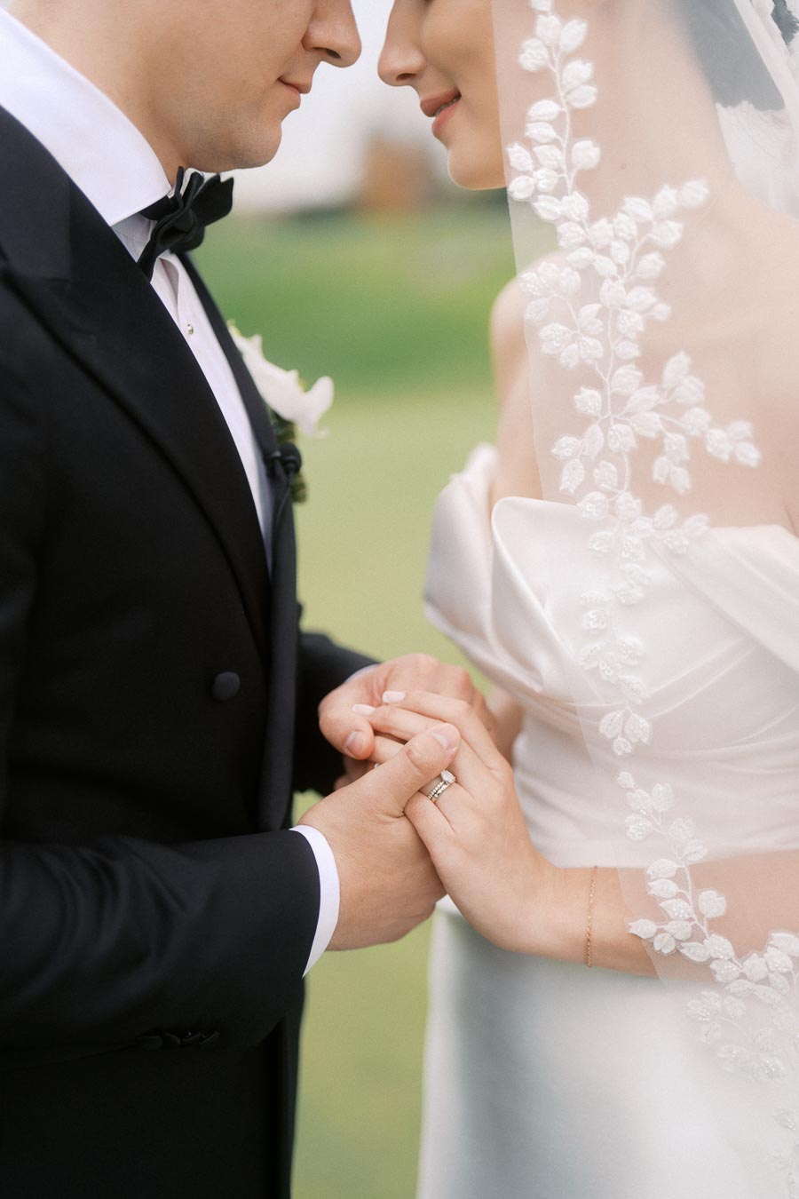 Close-up of a bride and groom holding hands during their wedding, with the bride wearing a lace veil and the groom in a black tuxedo.