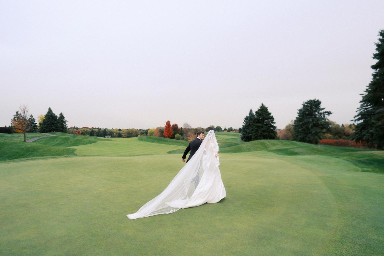 Bride and groom walking across a golf course, with the bride's long veil flowing behind them on a clear autumn day, surrounded by vibrant trees and green fairway.