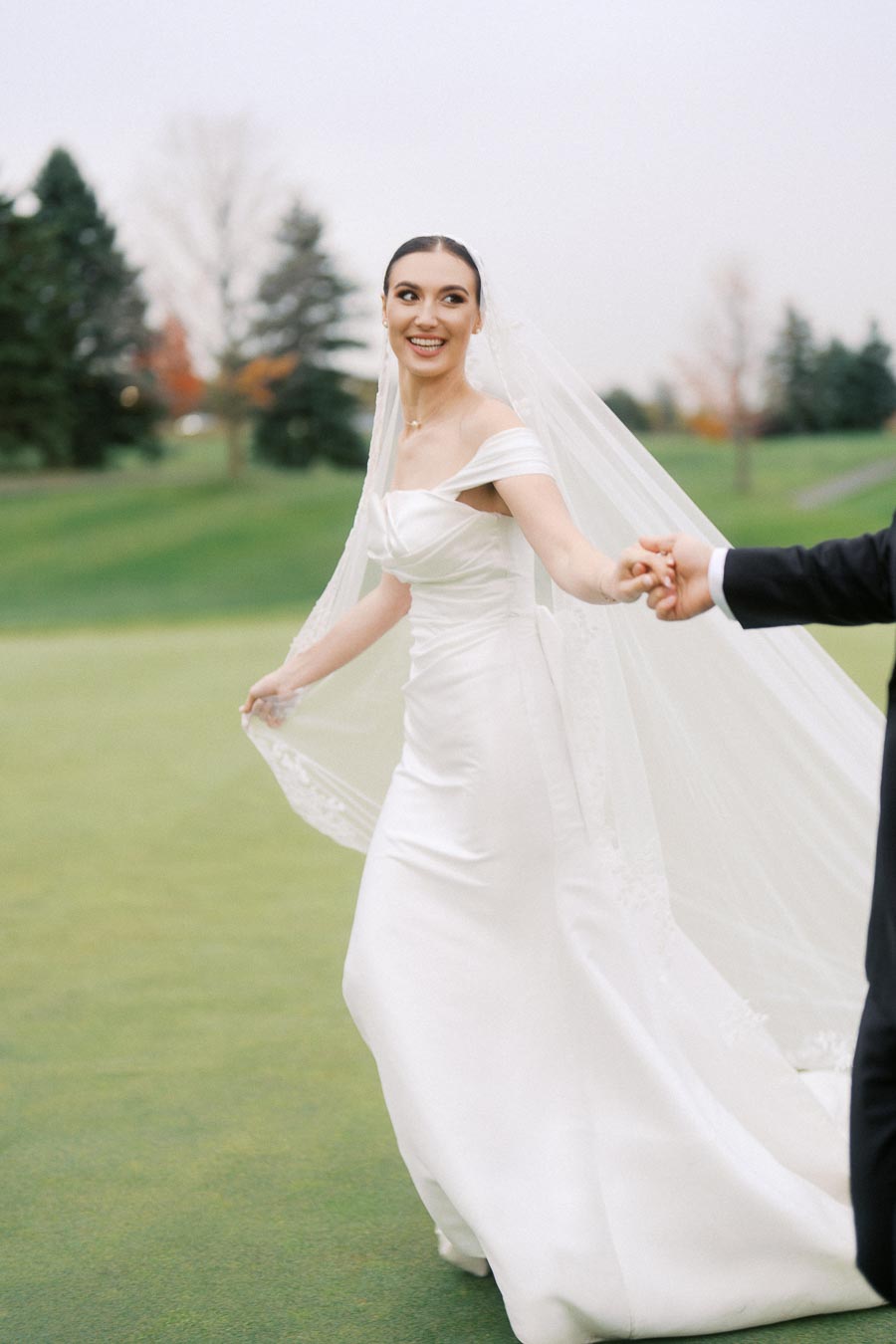 Bride in elegant white wedding dress and veil holding groom's hand in a serene outdoor setting with greenery.