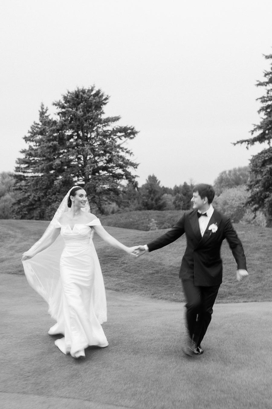 Black and white photo of a bride and groom joyfully holding hands while walking on a grassy landscape with trees in the background, capturing a candid wedding moment.