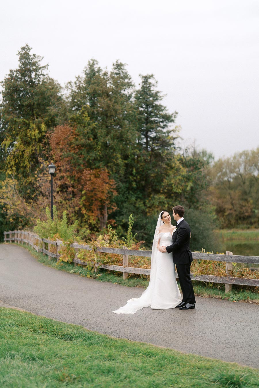 A bride and groom embrace on a scenic path surrounded by trees with autumn foliage, creating a romantic outdoor wedding scene.