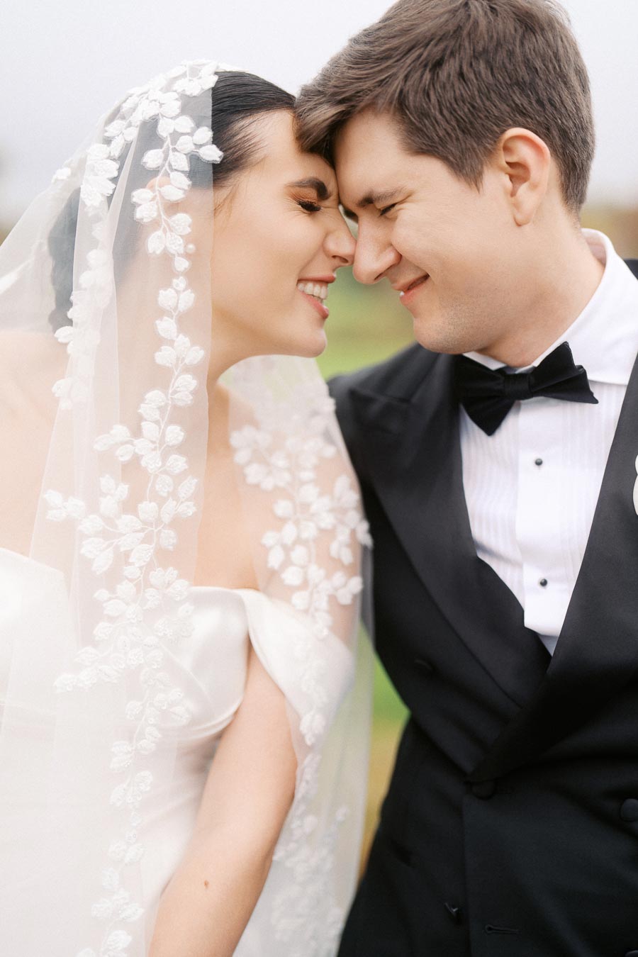 A joyful bride and groom share a tender moment during their outdoor wedding. The bride wears a veil adorned with floral lace, and the groom is dressed in a classic black tuxedo with a bow tie.