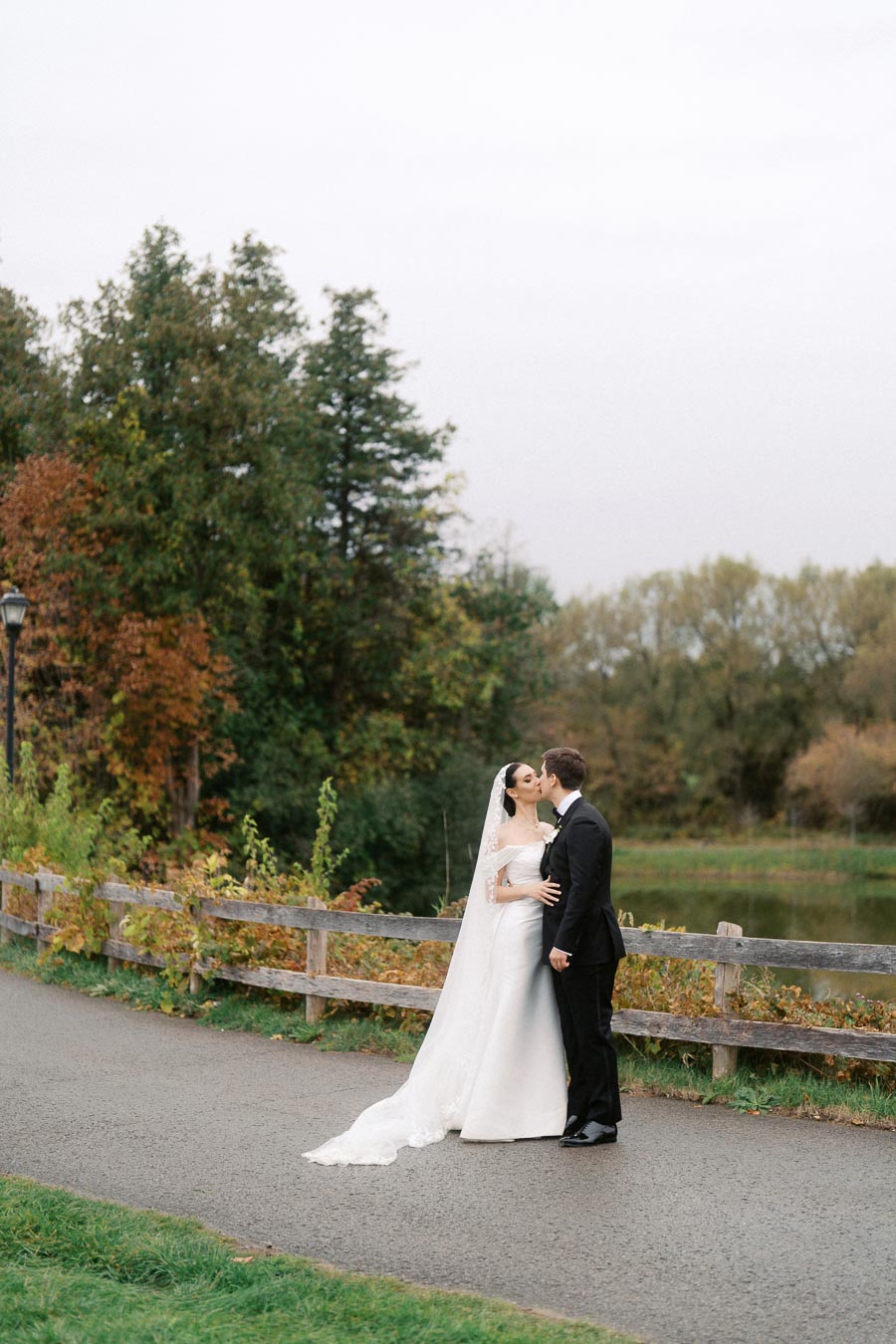Elegant couple shares a romantic kiss during an outdoor wedding photoshoot by a picturesque lakeside with lush greenery and autumn foliage.