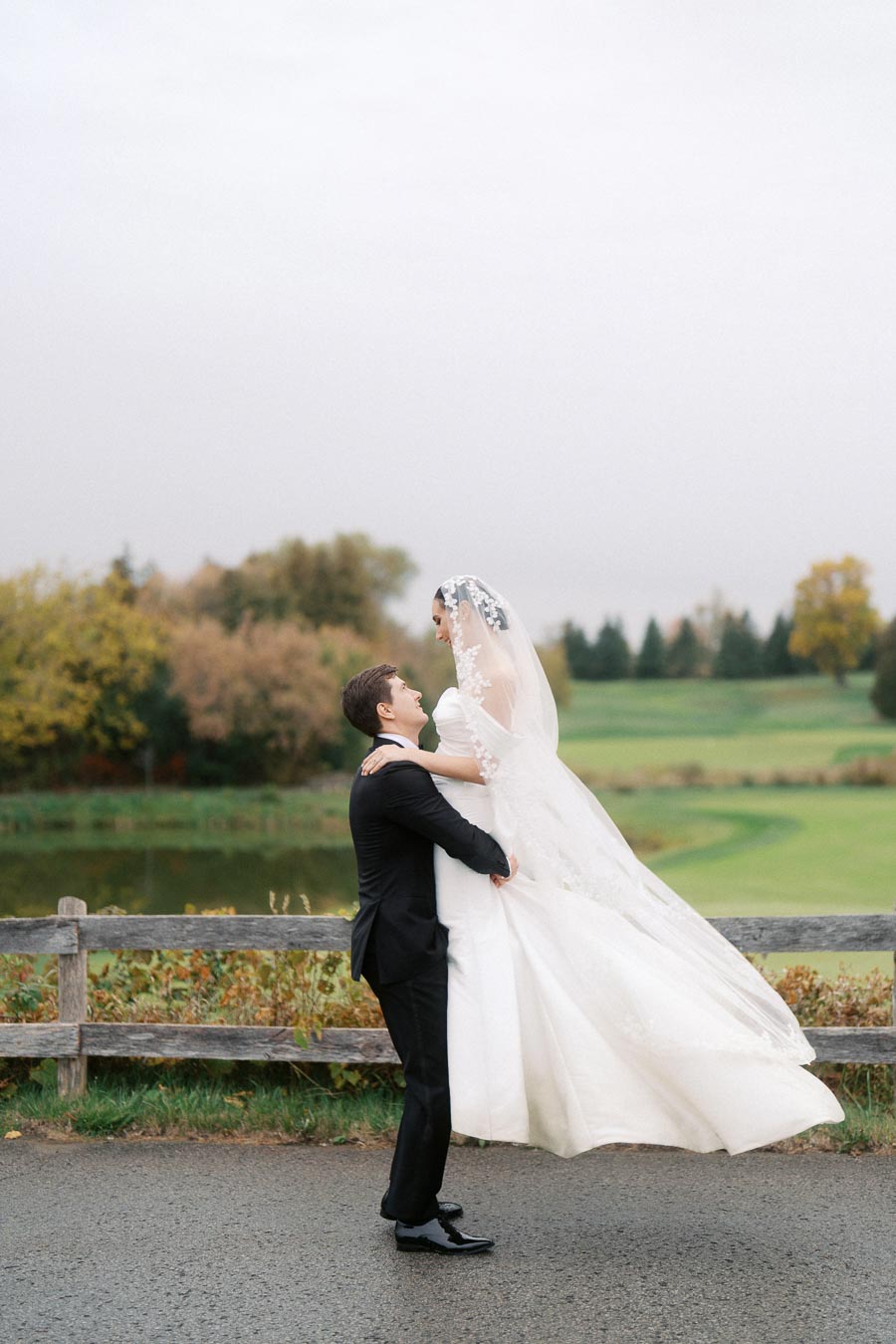 Bride in elegant white wedding dress lifted by groom in a tuxedo, smiling in a picturesque outdoor setting with lush greenery and wooden fence in the background.