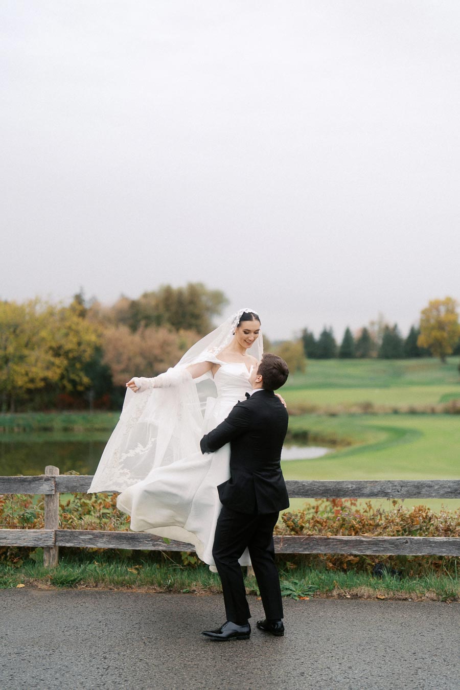 Bride and groom celebrating outdoors on their wedding day, with the groom lifting the bride joyfully near a rustic wooden fence and a scenic landscape in the background.