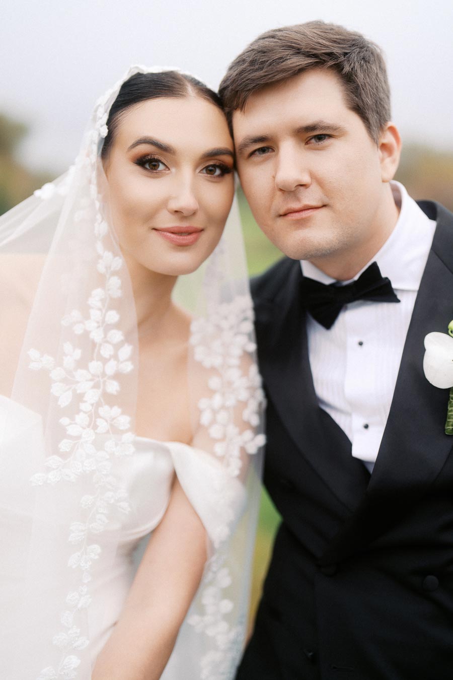 A newlywed couple posing together, with the bride wearing an elegant lace veil and the groom in a classic black tuxedo, capturing a timeless wedding moment.