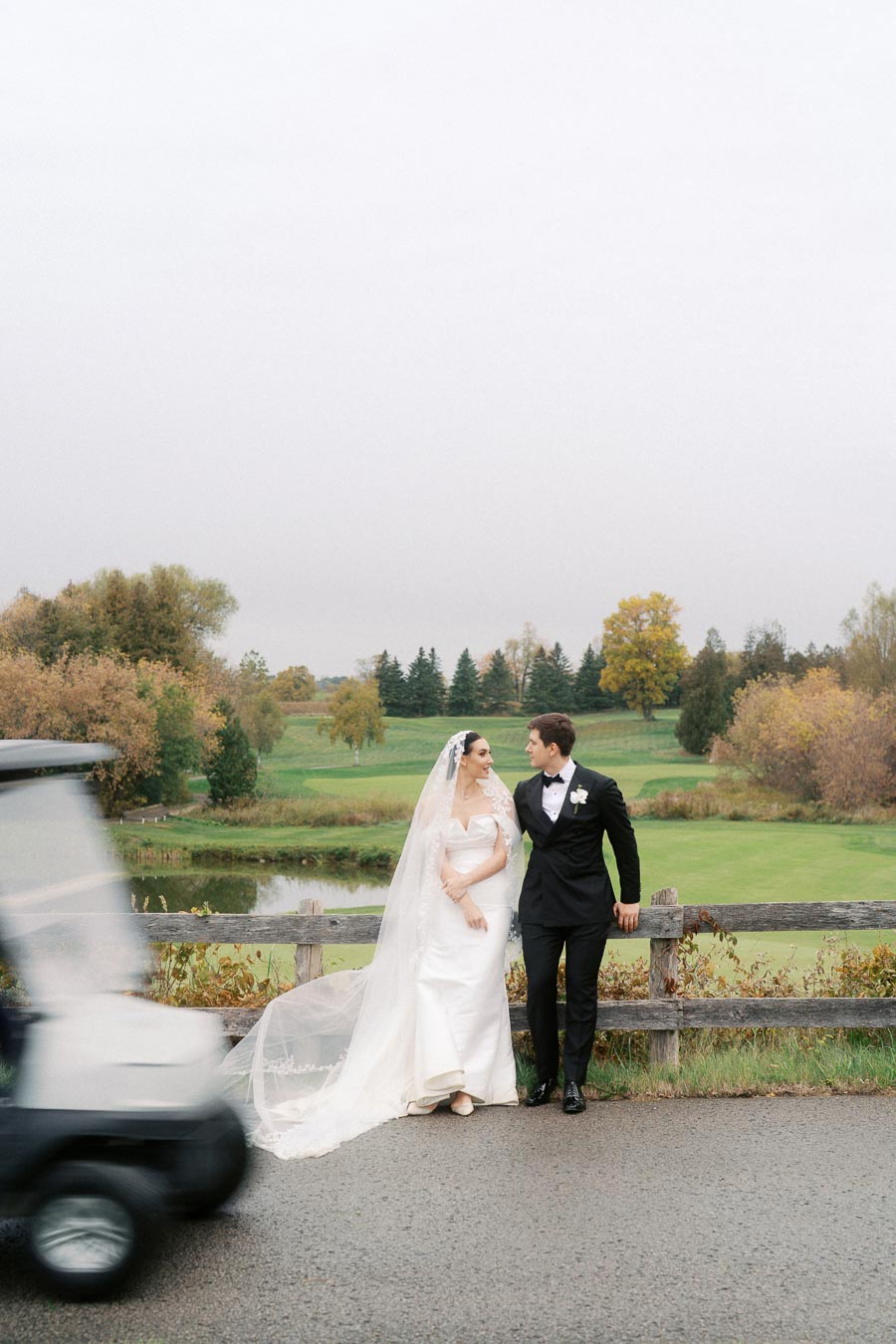 Bride and groom in elegant wedding attire stand by a wooden fence, set against a picturesque golf course with autumn foliage, as a golf cart passes by.