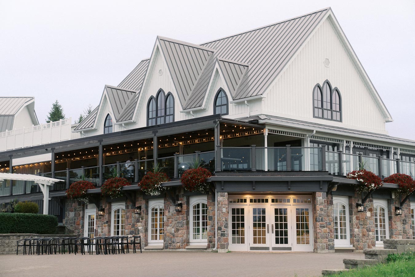 Modern rustic event venue with stone and white wood exterior, featuring large arched windows and a wraparound balcony adorned with vibrant flower arrangements. The building is set against a serene backdrop of lush greenery, creating a picturesque setting.