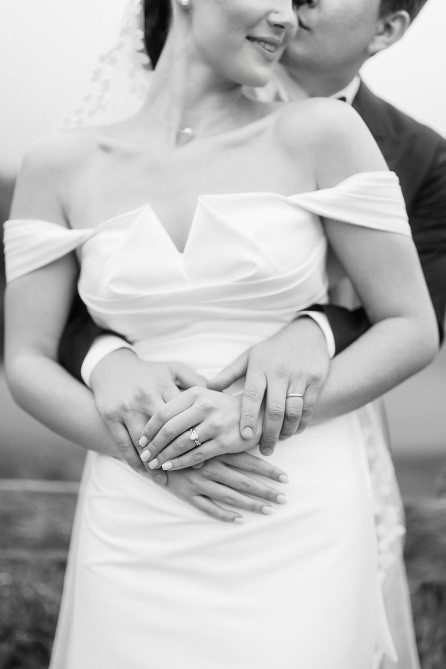 Black and white photo of a bride and groom embracing, highlighting their hands with wedding rings and the bride's elegant white gown.
