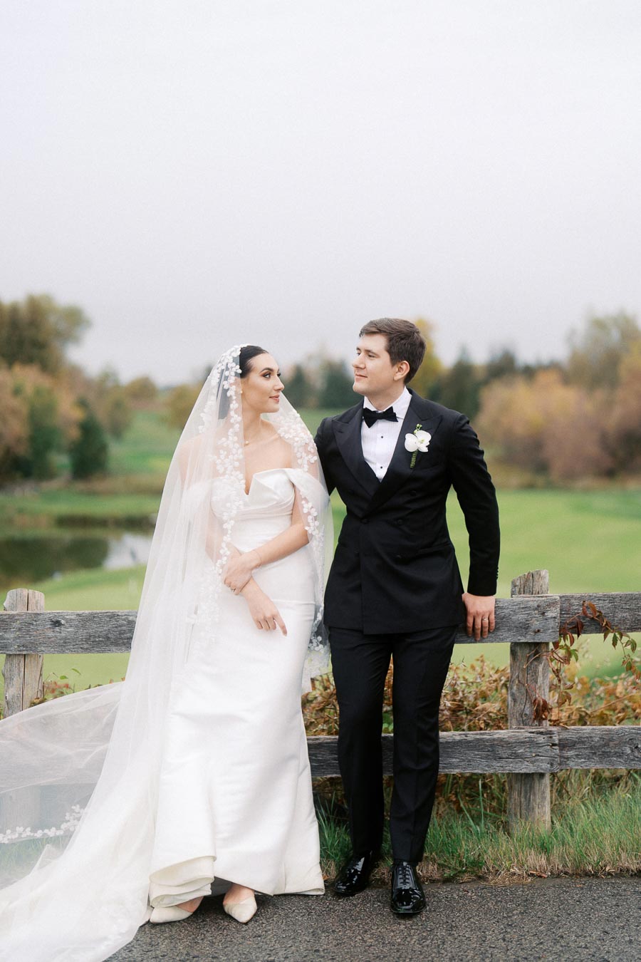 Elegant bride in white gown and veil stands next to groom in black tuxedo in scenic outdoor setting with wooden fence and lush greenery, capturing a classic wedding moment.