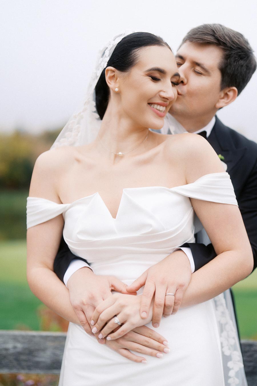 Smiling bride in elegant white gown embraced by her groom in a black suit during an outdoor wedding photoshoot.