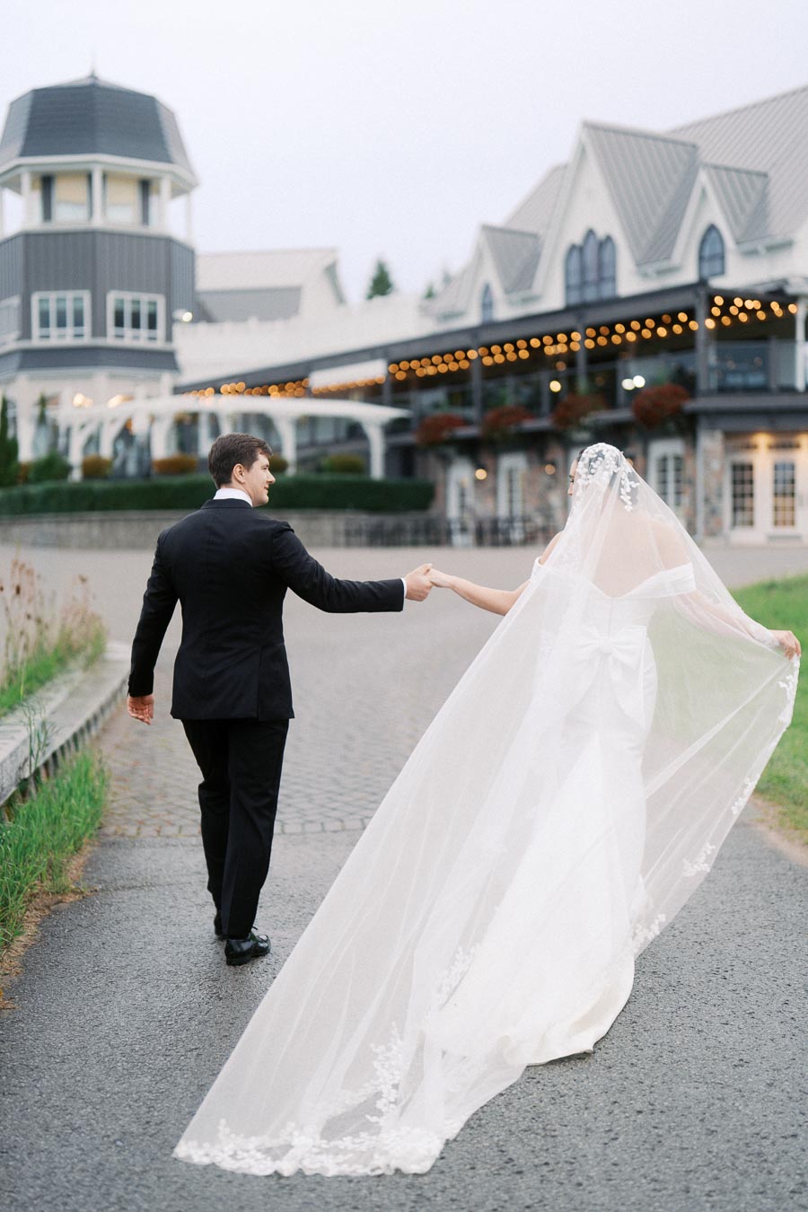 Bride and groom walking hand in hand in front of a charming venue with decorative lights.