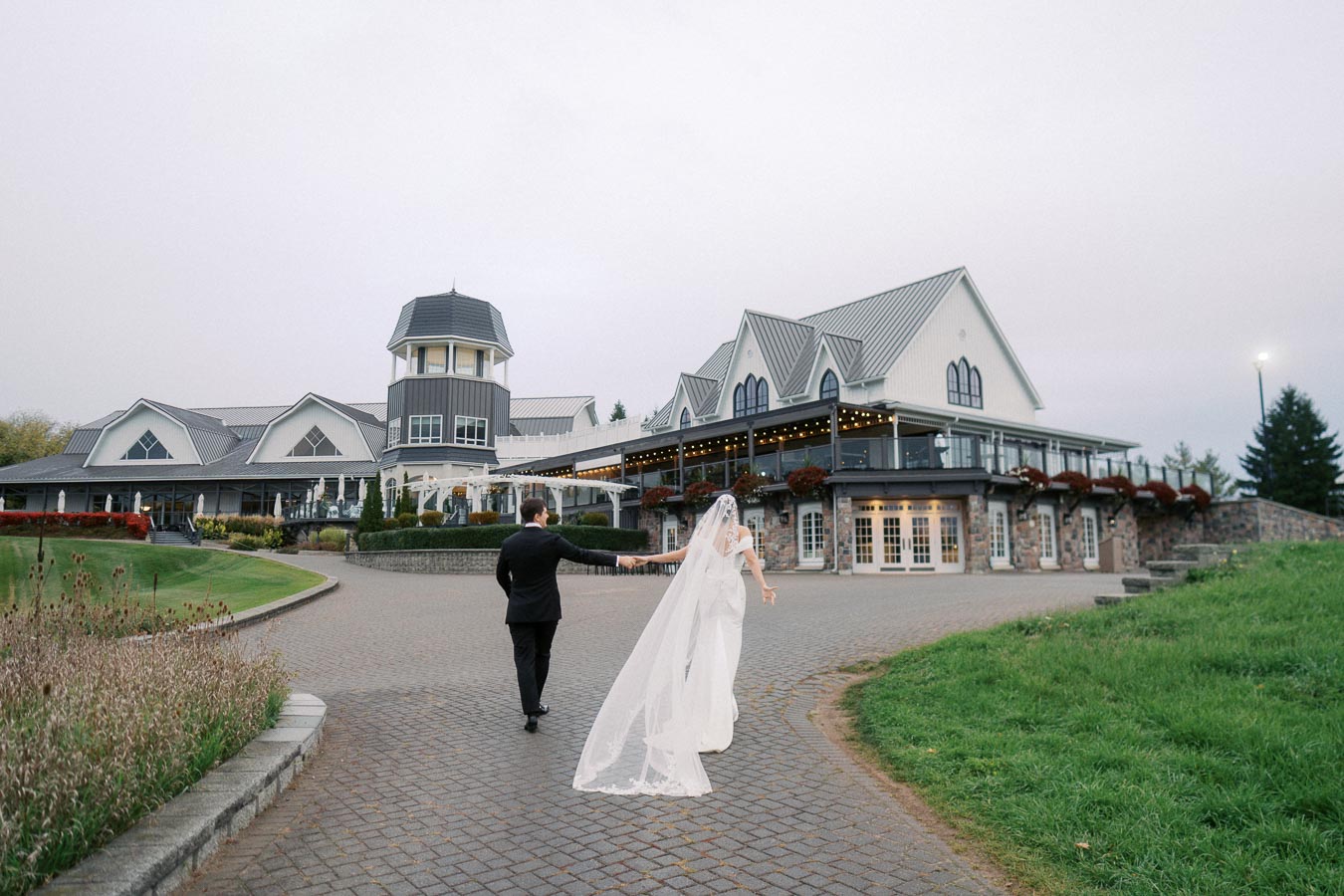 Elegant bride and groom walking hand in hand on a stone path towards a grand wedding venue with Victorian-style architecture, surrounded by lush greenery and overcast sky.