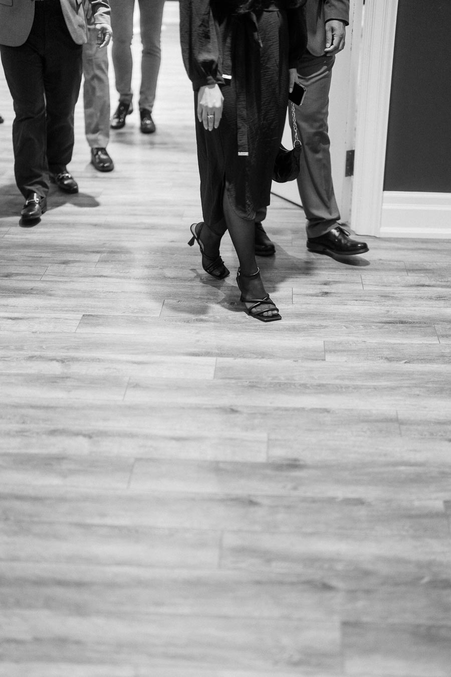 Black and white image of a group of people walking on a wooden floor in formal attire, emphasizing the lower half of the body, with focus on elegant shoes and clothing.