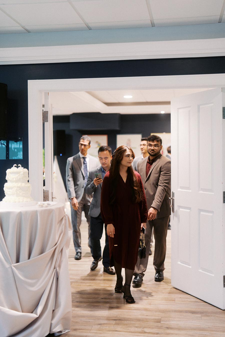 Guests entering a wedding reception hall, with a white tiered cake on a table, dressed in formal attire.