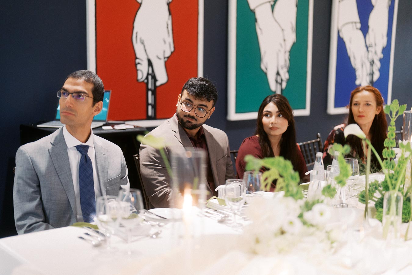 A group of four people in formal attire sitting at a decorated table during a meeting or event, with colorful artwork in the background.