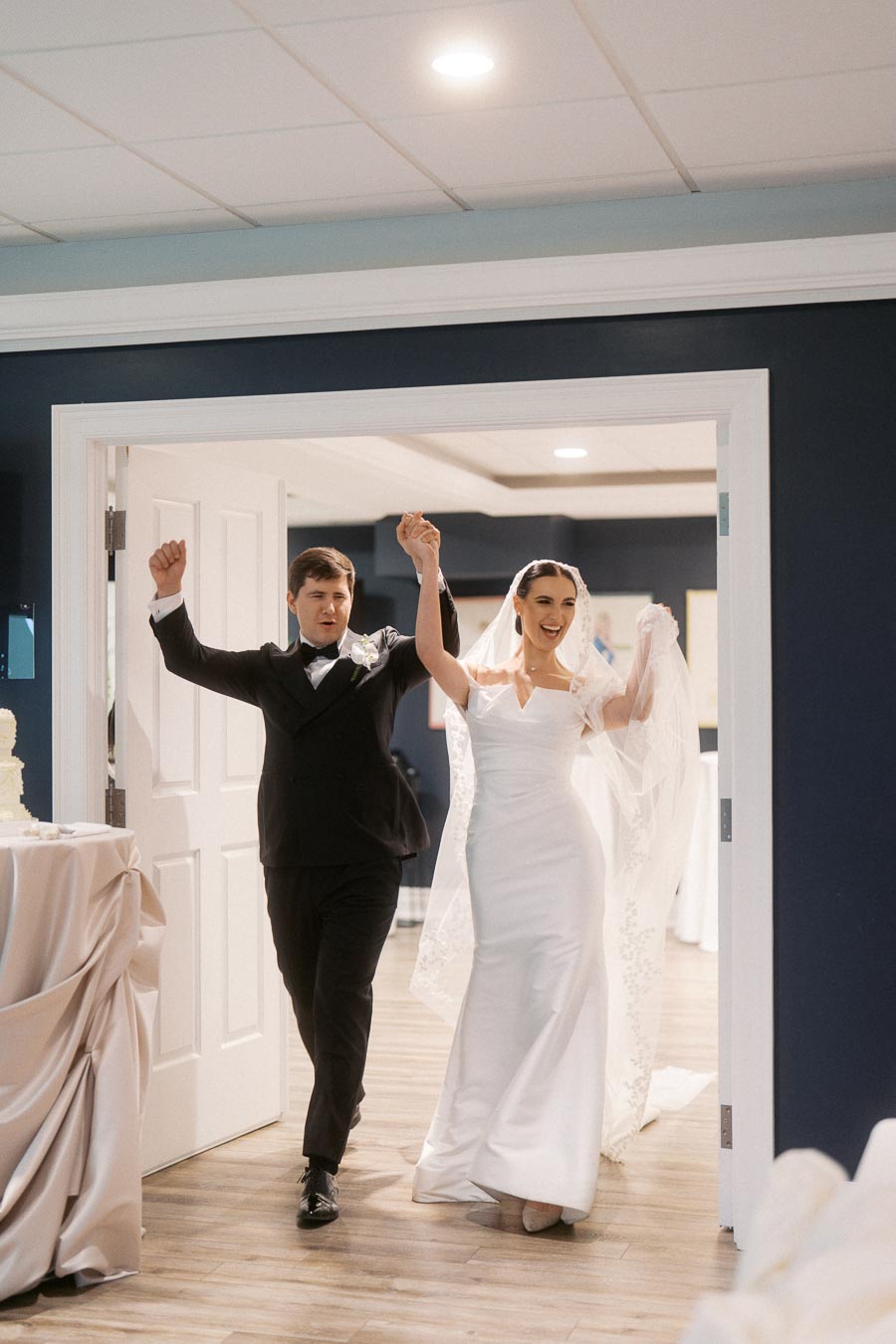Happy newlywed couple joyfully entering a wedding reception, with the bride in a stunning white gown and veil, and the groom in a stylish black tuxedo, both raising their arms in celebration.