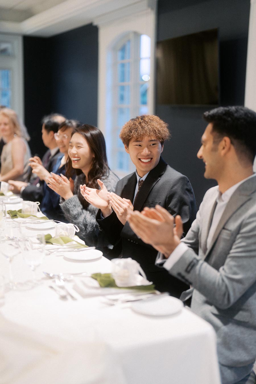 Group of people in formal attire sitting and clapping at an elegant dinner table, expressing joy and celebration at a social event or gathering in a well-lit room.