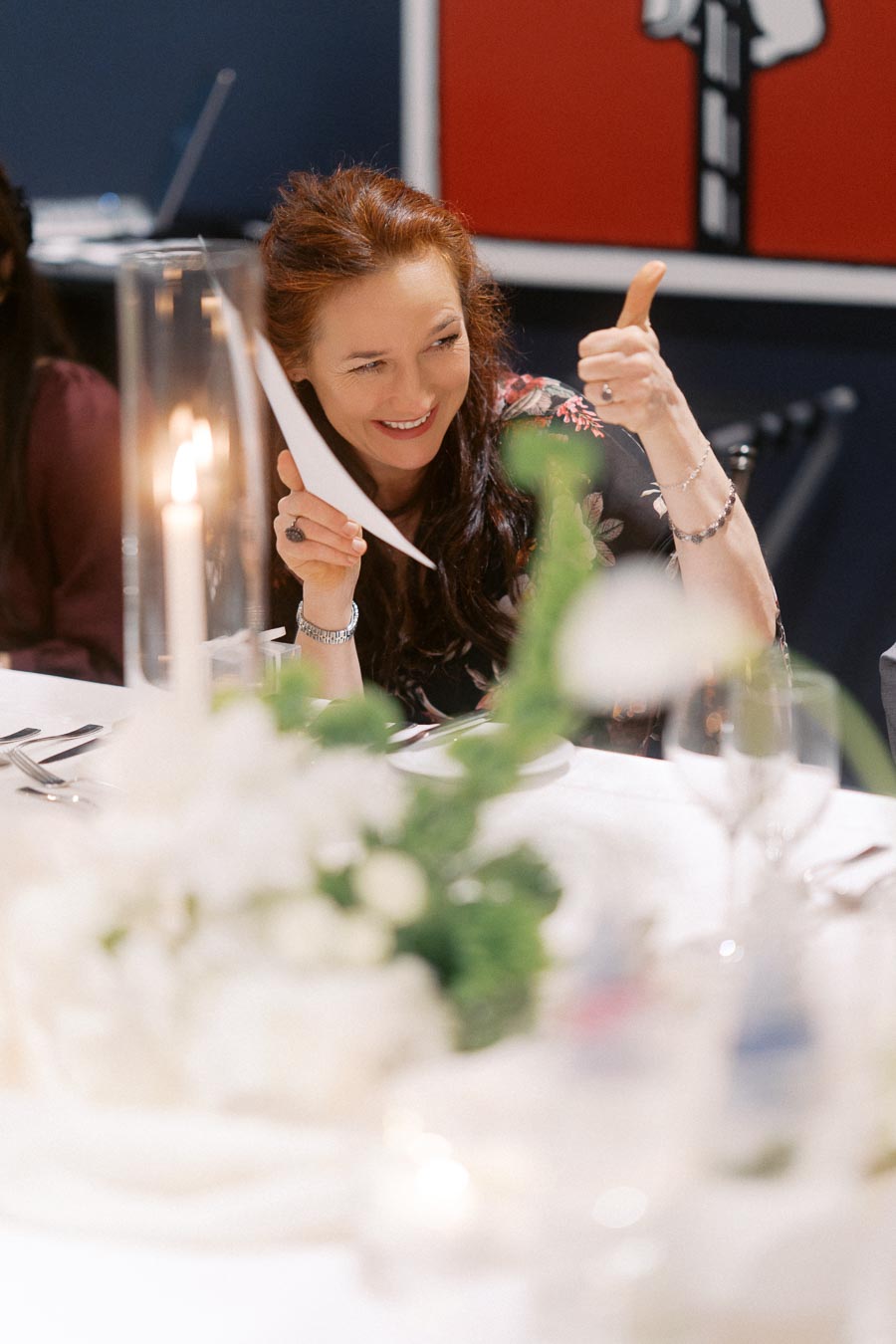 Woman smiling and giving a thumbs up at a dining table with elegant decor, including flowers, candles, and glassware.