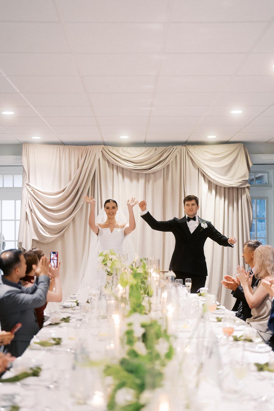 Bride and groom joyfully entering the reception hall, celebrating their wedding day with guests clapping and taking photos, elegant wedding decor with white and green floral centerpiece.