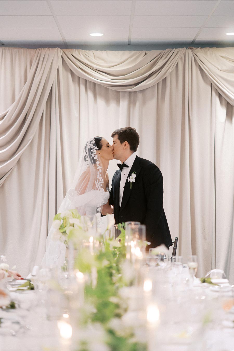 Bride and groom sharing a romantic kiss at their elegantly decorated wedding reception, with a backdrop of draped curtains and a beautifully set table adorned with flowers and candles.